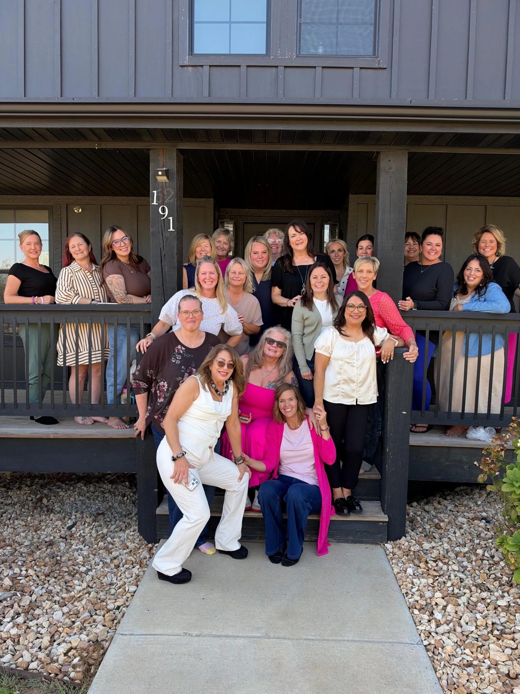 A large group of smiling women from The Boss Suite community posing together on the porch of a dark wooden house