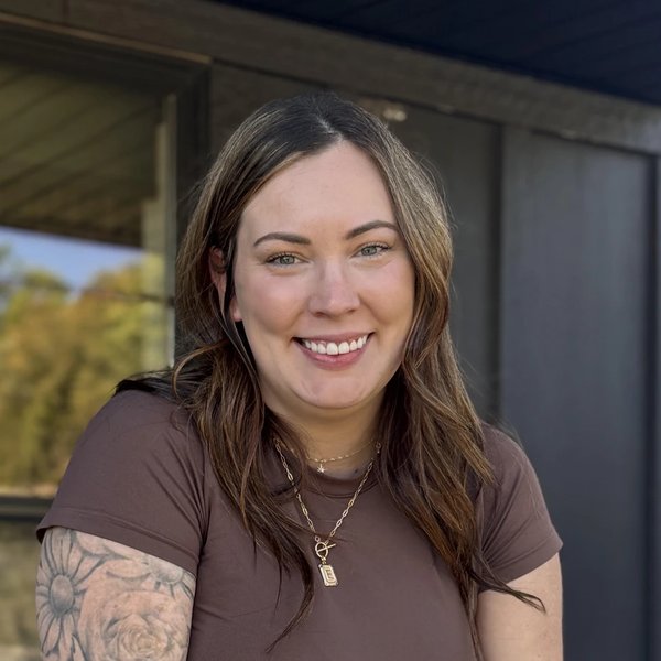 Harley smiling while leaning on a porch railing, wearing a brown t-shirt and gold necklace