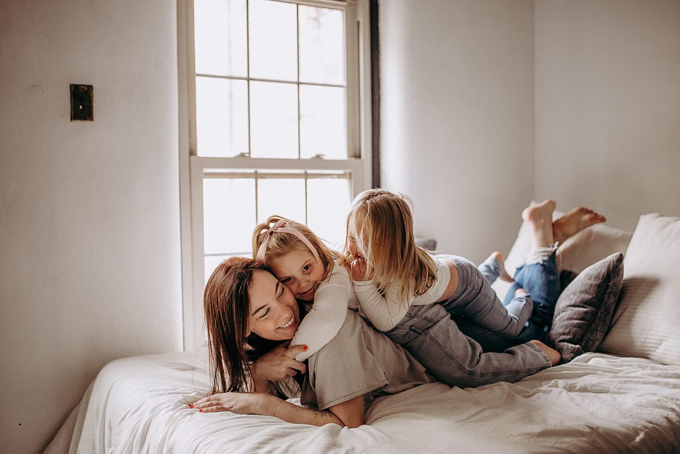 Harley smiling while lying on a bed by a sunny window as her two daughters pile onto her back